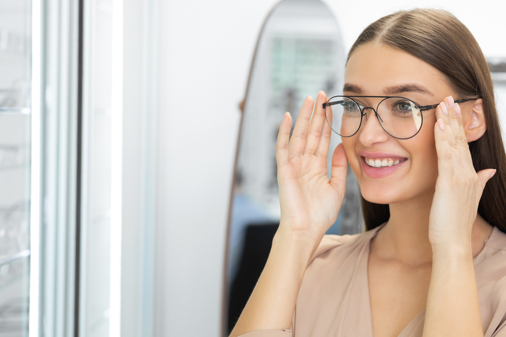 Smiling woman adjusting her eyeglasses while looking in a mirror, symbolizing proper fit, visual clarity, and comprehensive eye care services – eye care pearland tx