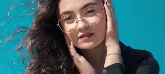 Stylish Clear Eyewear - Webster Eye Doctor | Clear Lake Eye Center A young woman with wavy brown hair wears clear-framed glasses and poses against a bright blue background. - Webster Eye Doctor | Clear Lake Eye Center