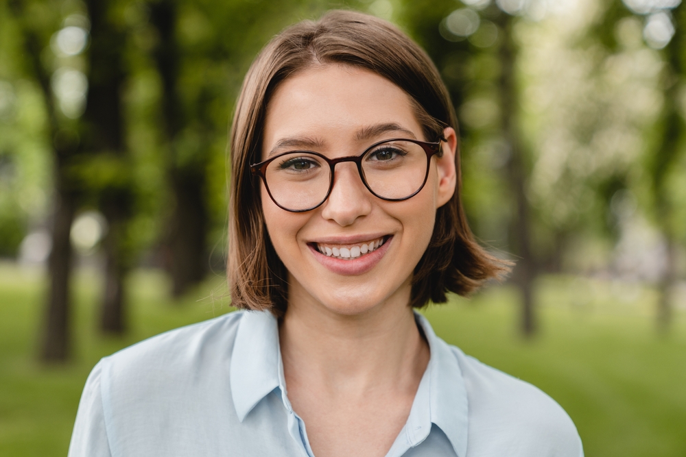 Smiling woman wearing eyeglasses outdoors representing clear vision and confidence – eye doctor pasadena tx Happy woman wearing round eyeglasses and smiling in a green outdoor setting, symbolizing comfortable vision correction and trusted eye care – eye doctor pasadena tx
