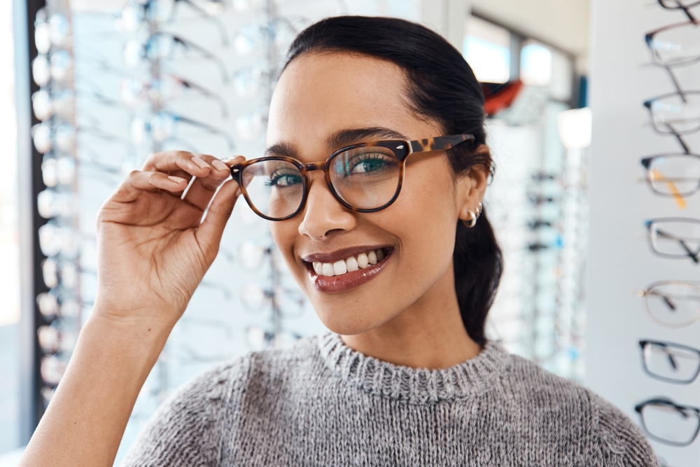 Happy woman adjusting her eyeglasses in an optical store setting, symbolizing proper fit, clarity, and personalized eye care services – eye care pearland tx