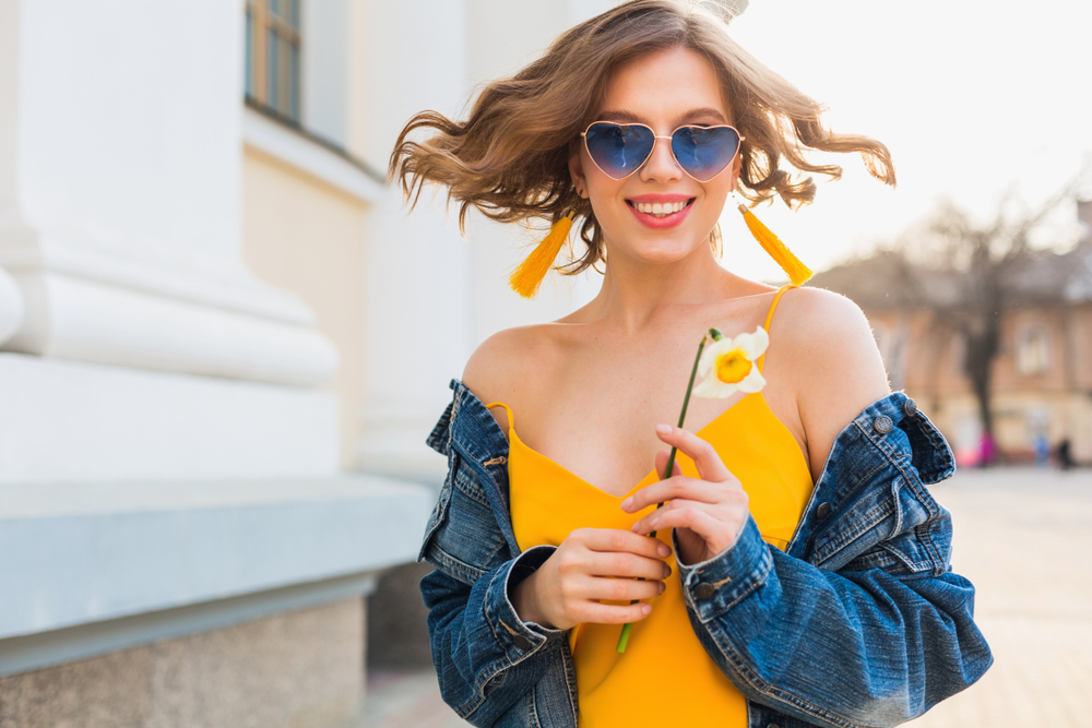 Woman wearing stylish sunglasses and smiling outdoors representing confident vision care – eye doctor pasadena tx Smiling woman wearing heart-shaped sunglasses and holding a flower in a sunlit outdoor setting, symbolizing fashionable eyewear and comprehensive eye care – eye doctor pasadena tx