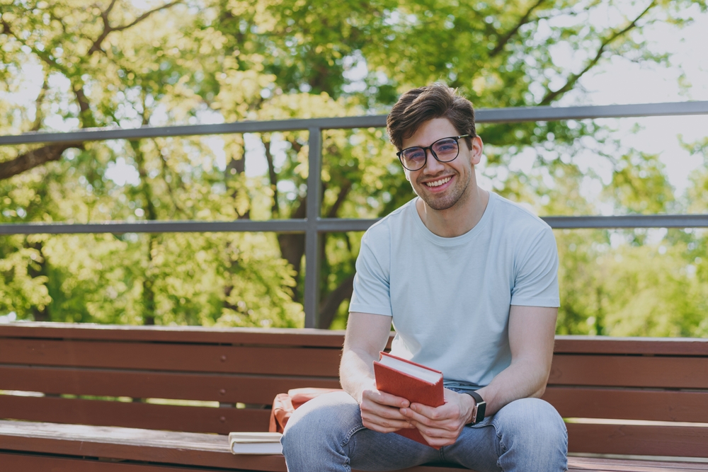 Clear Vision for Outdoor Reading - Webster Eye Doctor | Clear Lake Eye Center A young man wearing stylish glasses sits on a park bench, smiling while holding a book. - Webster Eye Doctor | Clear Lake Eye Center