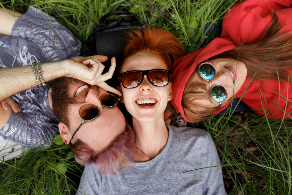 Stylish Shades and Smiles - Webster Eye Doctor | Clear Lake Eye Center Three friends wearing stylish sunglasses lie on the grass, laughing and enjoying a sunny day. - Webster Eye Doctor | Clear Lake Eye Center